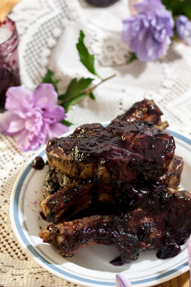 pile of pork ribs smothered in blueberry bbq sauce on white plate with purple flowers in background