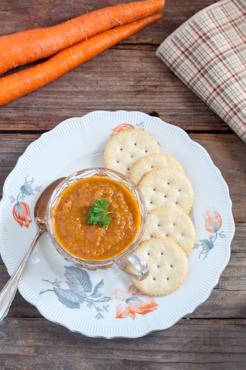 Glass cup on a white plate with orange roses filled with carrot soup and parsley garnish on a plate with crackers