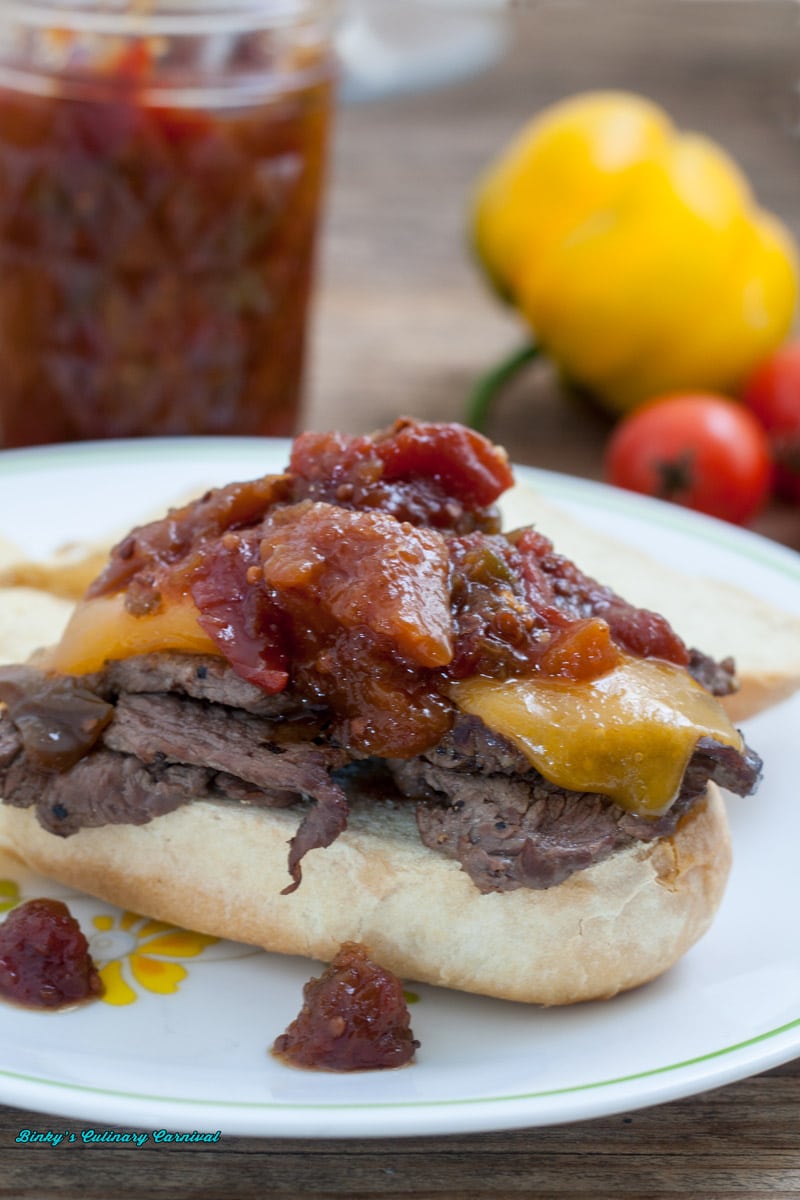 Close up of hoagie roll steak sandwich with melted cheddar topped with oscar relish on white plate