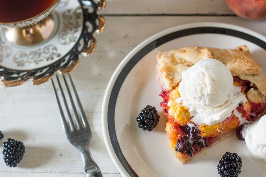 Piece of blackberry peach galette on on white plate with black rim, topped with a scoop of ice cream.