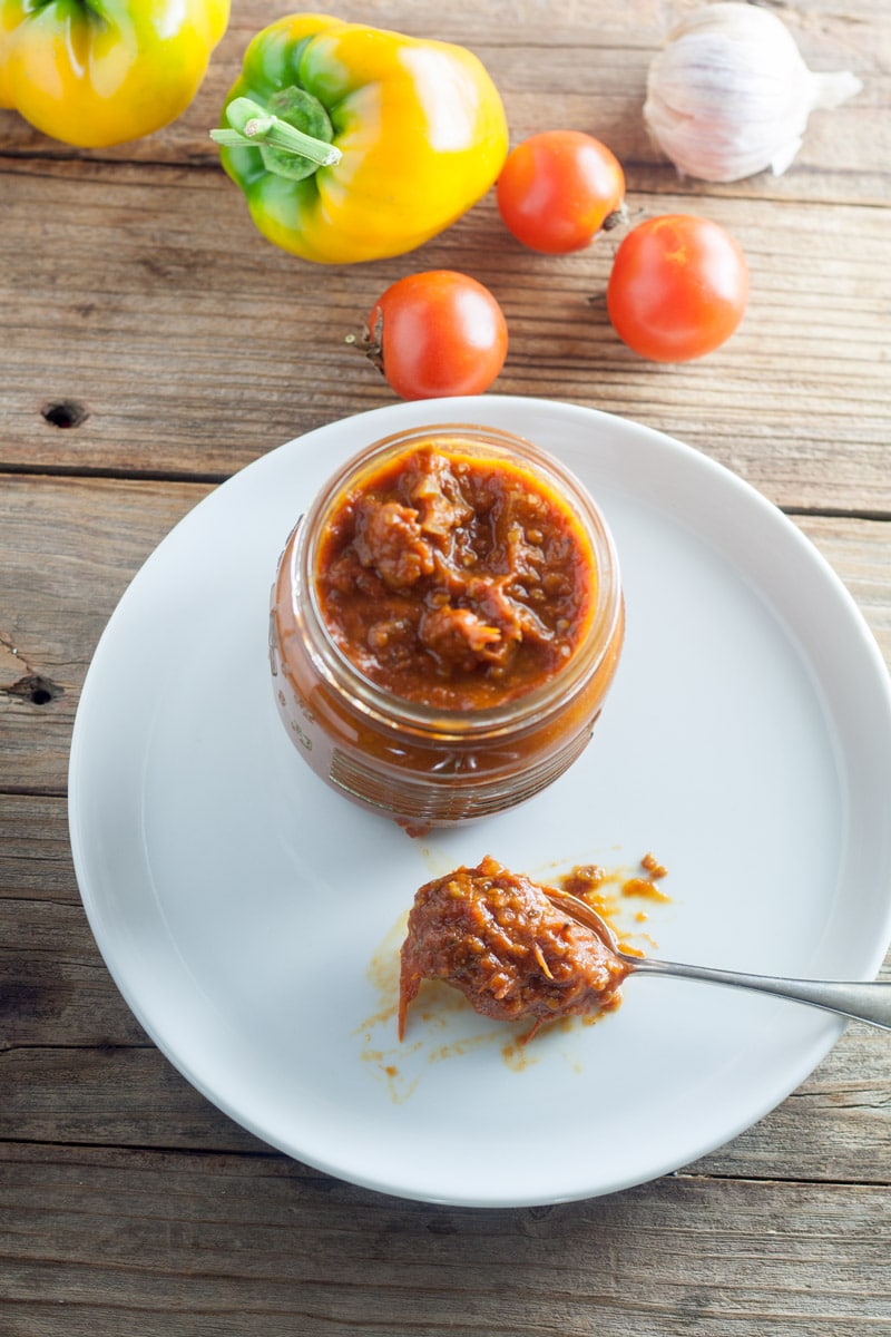 Silver spoon filled with salsa on white plate next to jar of canned salsa with cherry tomatoes, yellow peppers and garlic heads