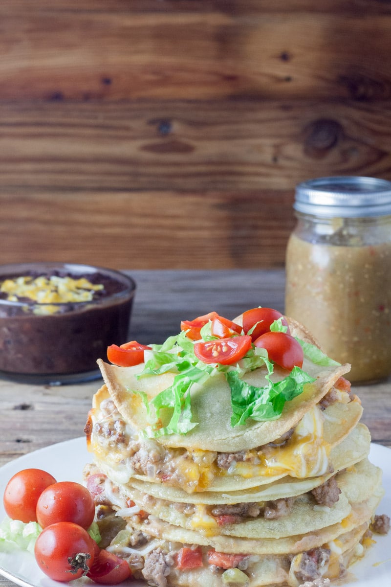 Corn tortillas stacked on top of each other with layers of beef and melted cheese, topped with tomatoes and lettuce on antique barn wood board.