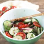 Sliced Cucumbers and Tomatoes in green bowl with red onion and a wood serving spoon