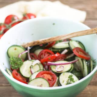 Sliced Cucumbers and Tomatoes in green bowl with red onion and a wood serving spoon