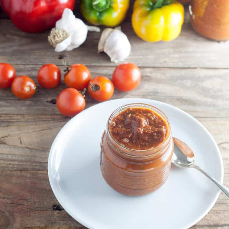 Jar of salsa with silver spoon on white plate with tomatoes, peppers, garlic and jars of salsa in the background