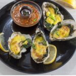Oyster on a black plate topped with Holladaise sauce and spinach. Oysters Rockefeller style.