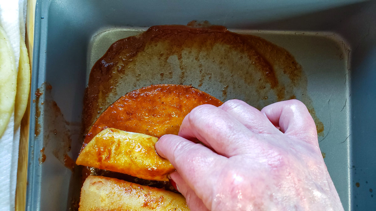 Dipped tortilla in enchilada sauce with women's hand rolling it up.