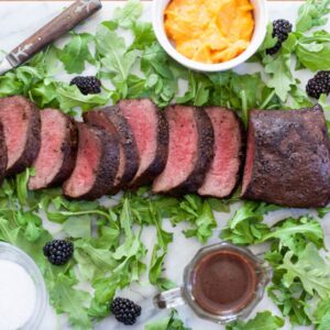 Medium rare sliced venison loin on a bed of arugula with strewn blackberries and sides in small bowls.