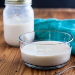 Clear glass bowl with evaporated milk with a jar and green tea towel in the background.