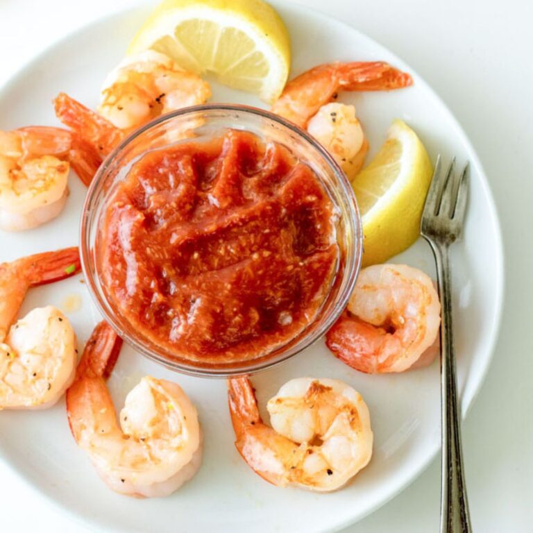 Garlic shrimp surrounding a bowl of homemade cocktail sauce on a white plate with lemons.