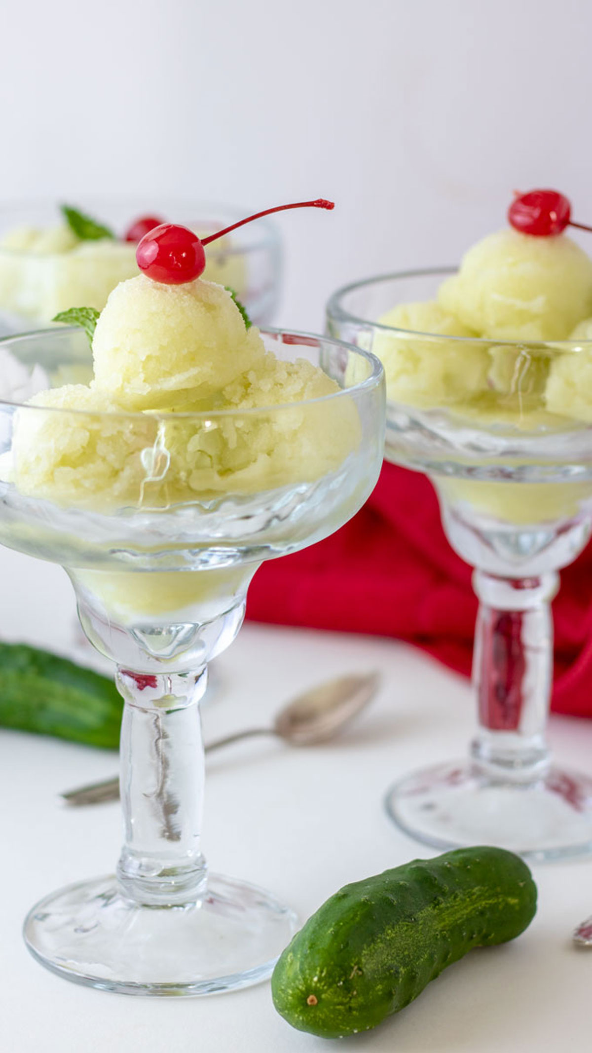 Cucumber sorbet in three glasses topped with cherry with red napkin and cucumbers.