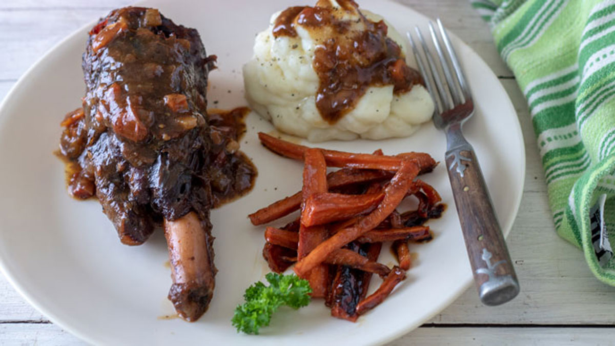 White plate with shank, potatoes, sauce and carrots.