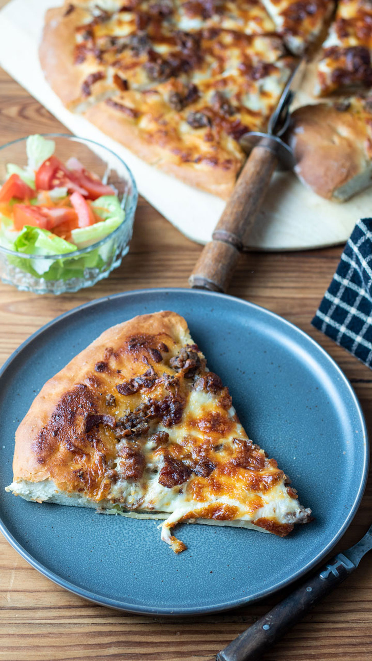 Slice of pizza on a blue plate next to a bowl of salad and a whole pie.