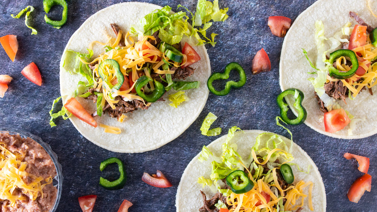 2 tostadas with shredde venison, shredded lettuce, diced tomatoes and shredded sharp cheddar cheese Overhead photo of tacos and refried beans.