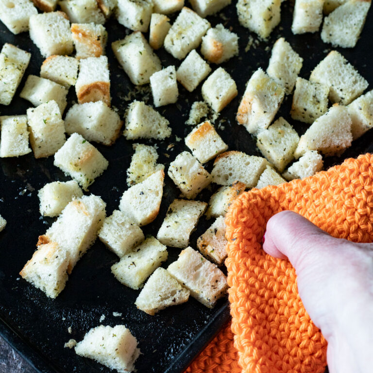 Woman's hand holding an orange hot pad grabbing a black tray of homemade croutons.