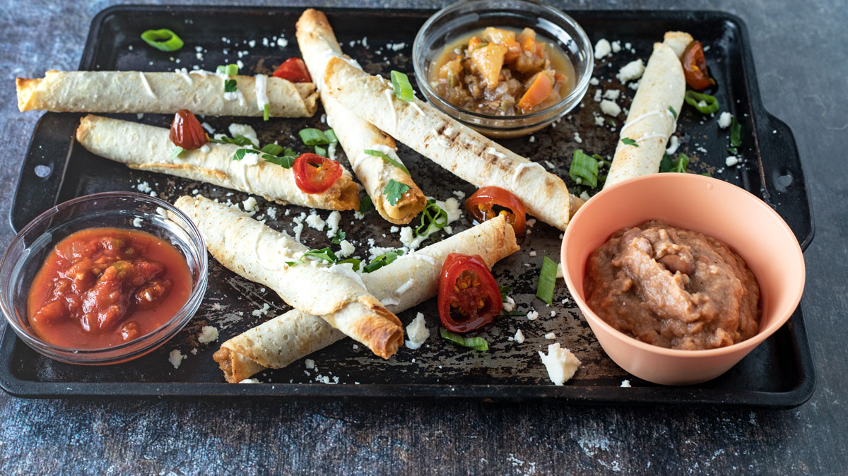 Air fryer taquitos arranged on board with crumbled cheese and green onions.