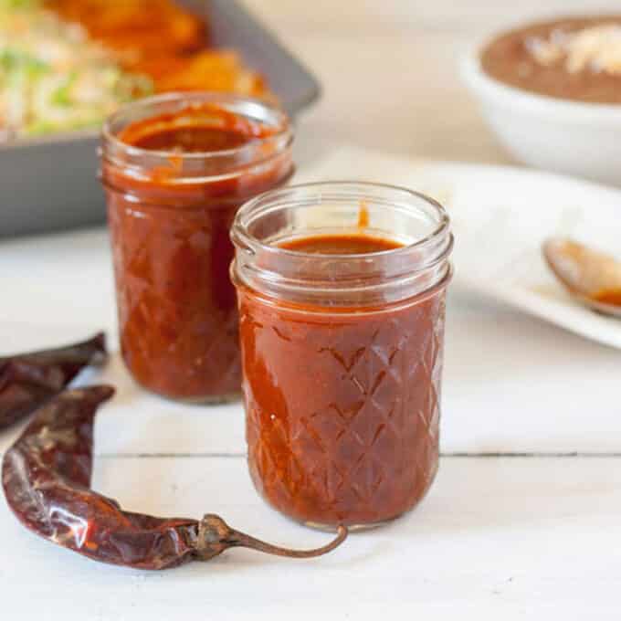 REd enchilada sauce in glass jars with tray of enchiladas in background.