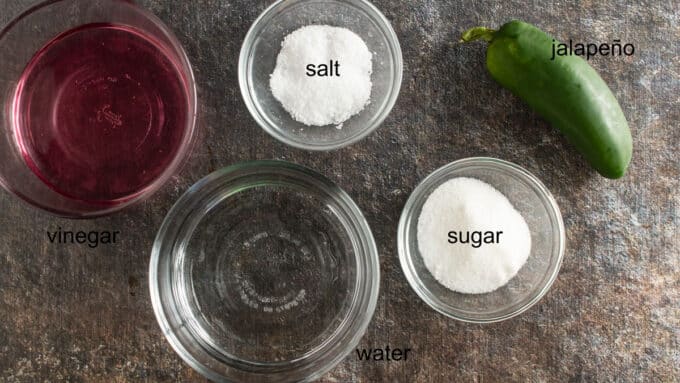 Salt, sugar, jalaeño, water and vinegar on dark board.