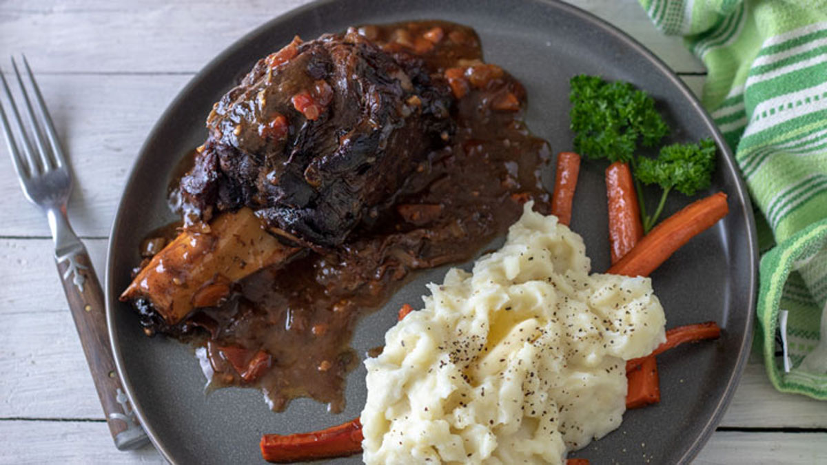 Shank covered in gravy on gray plate with veggies.