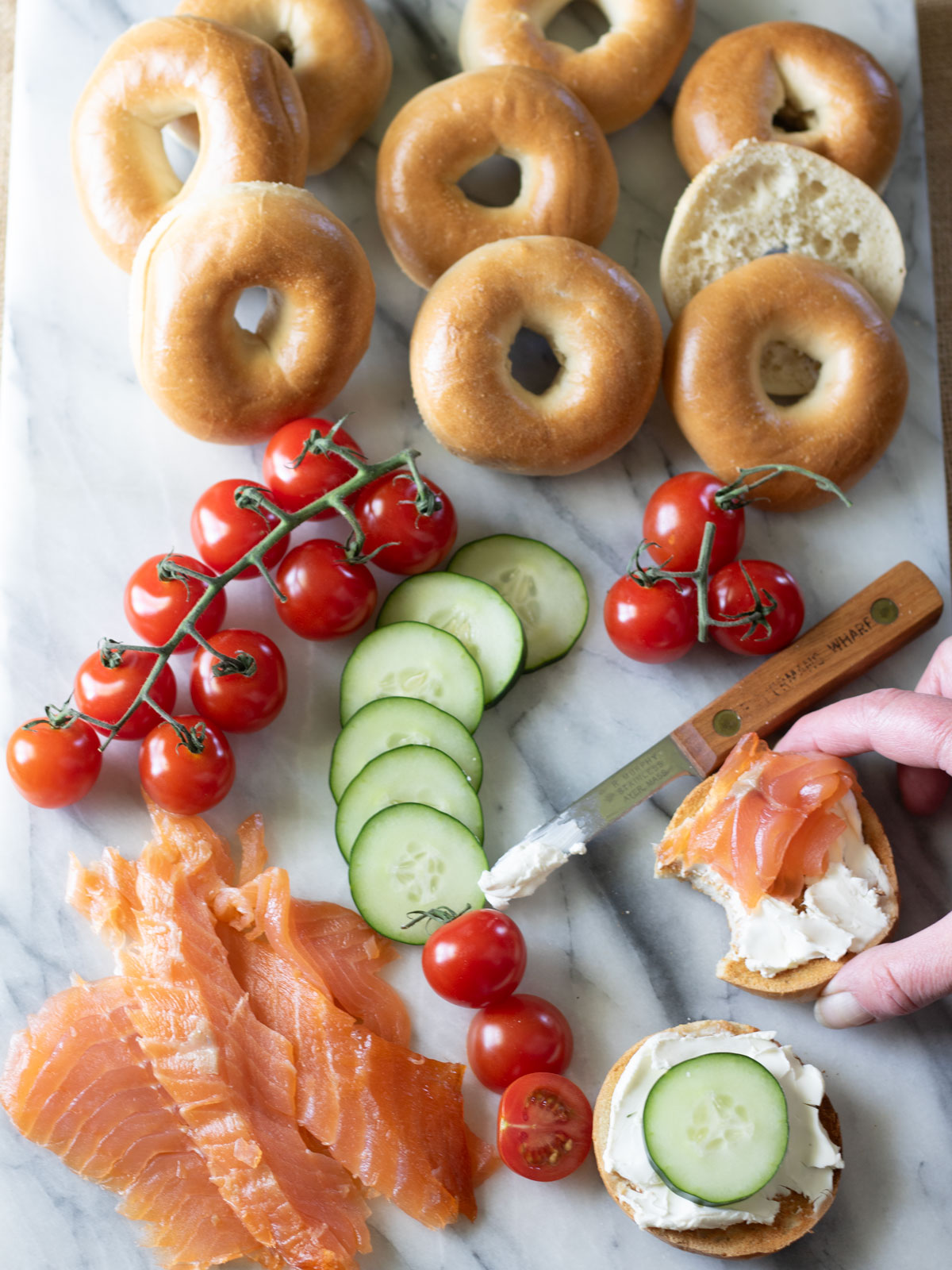 Hand grabbing bagel with lox. Sliced salmon on marble board with tomatoes, cucumbers and bagels.