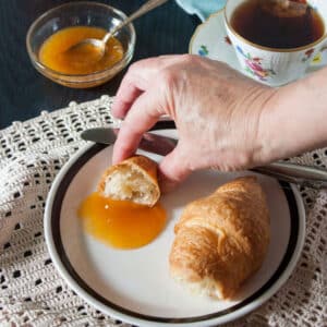 Womans hand scooping peach jam onto croissant.