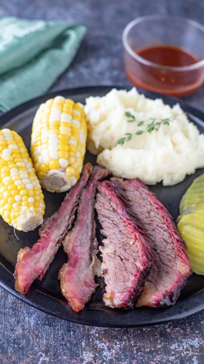 Slices of brisket with potatoes, corn on the cob and pickles.