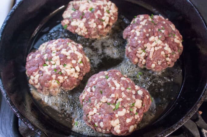 Four burgers frying in cast iron pan.