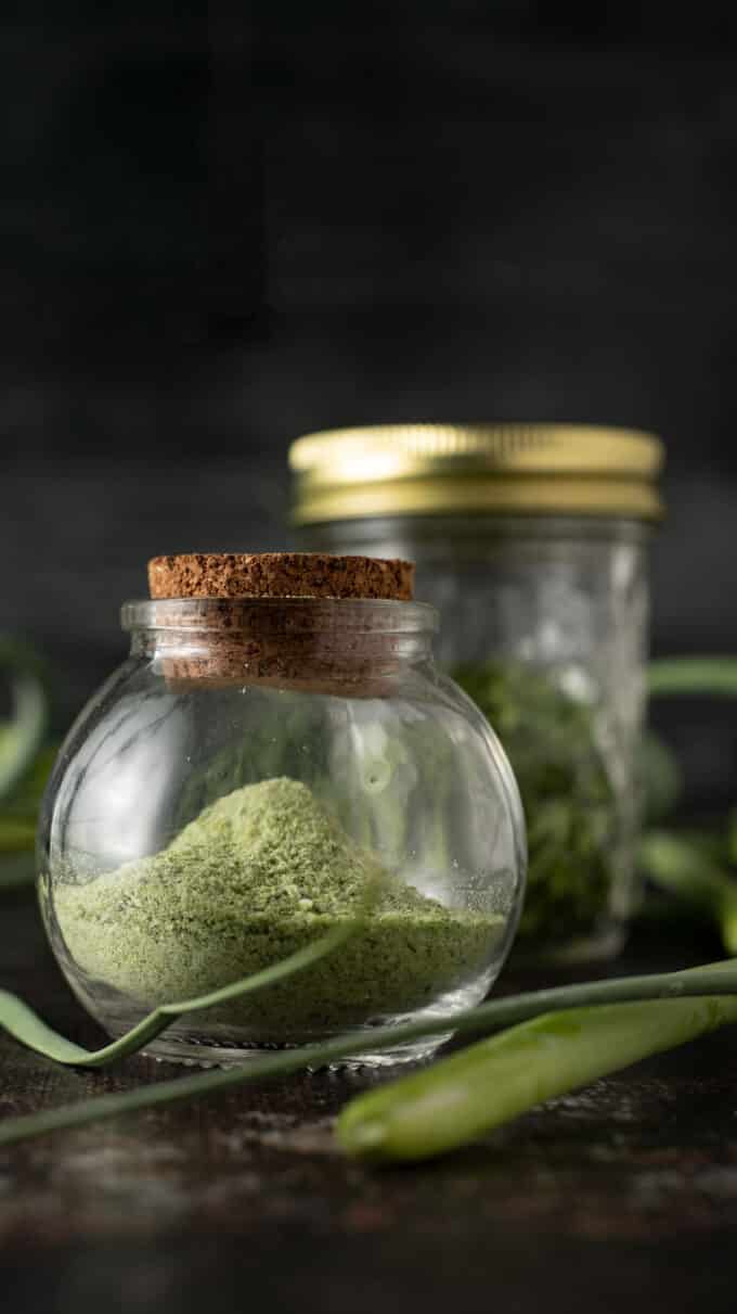 Small round jar of garlic scape powder with fresh garlic scapes around it.