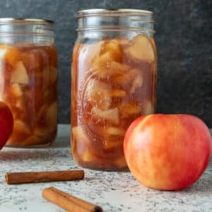 Jars of apple pie filling with fresh apples and cinnamon sticks.
