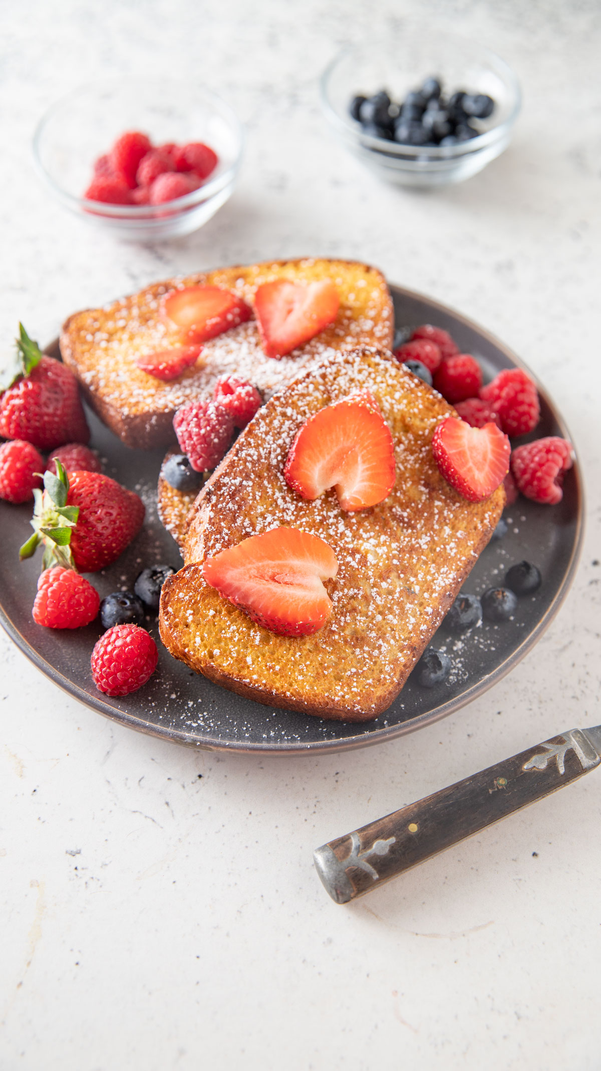 French toast on a gray plate with sliced strawberries on top and blueberry and raspberries..