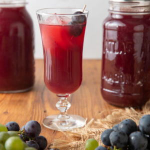 Grape juice in a fluted glass next to jars of canned Grape juice.