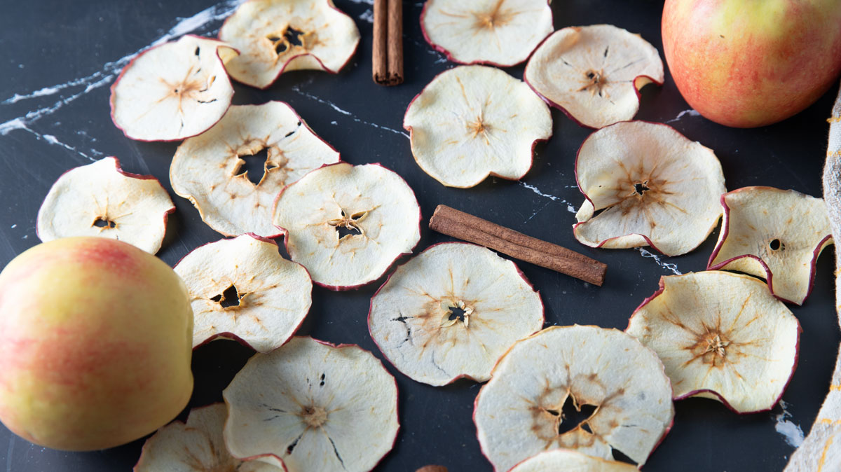 Dried apple slices scattered on a black board with cinnamon and fresh apples.
