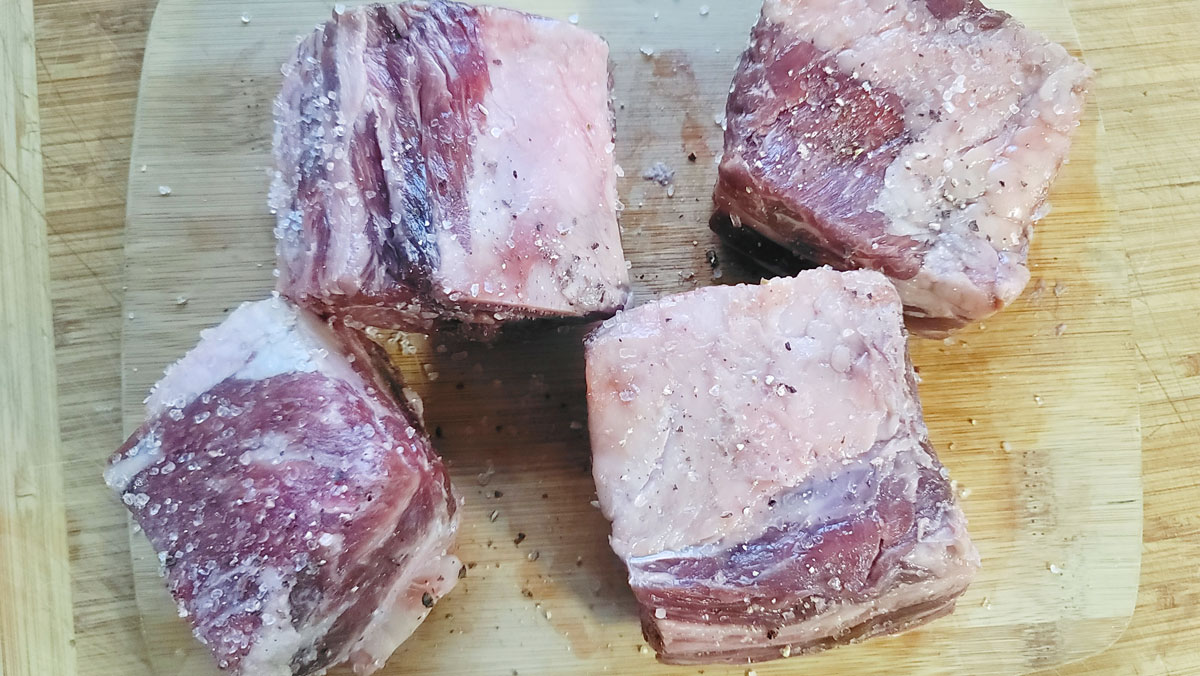 Beef short ribs on a cutting board after being seasoned with salt and pepper.