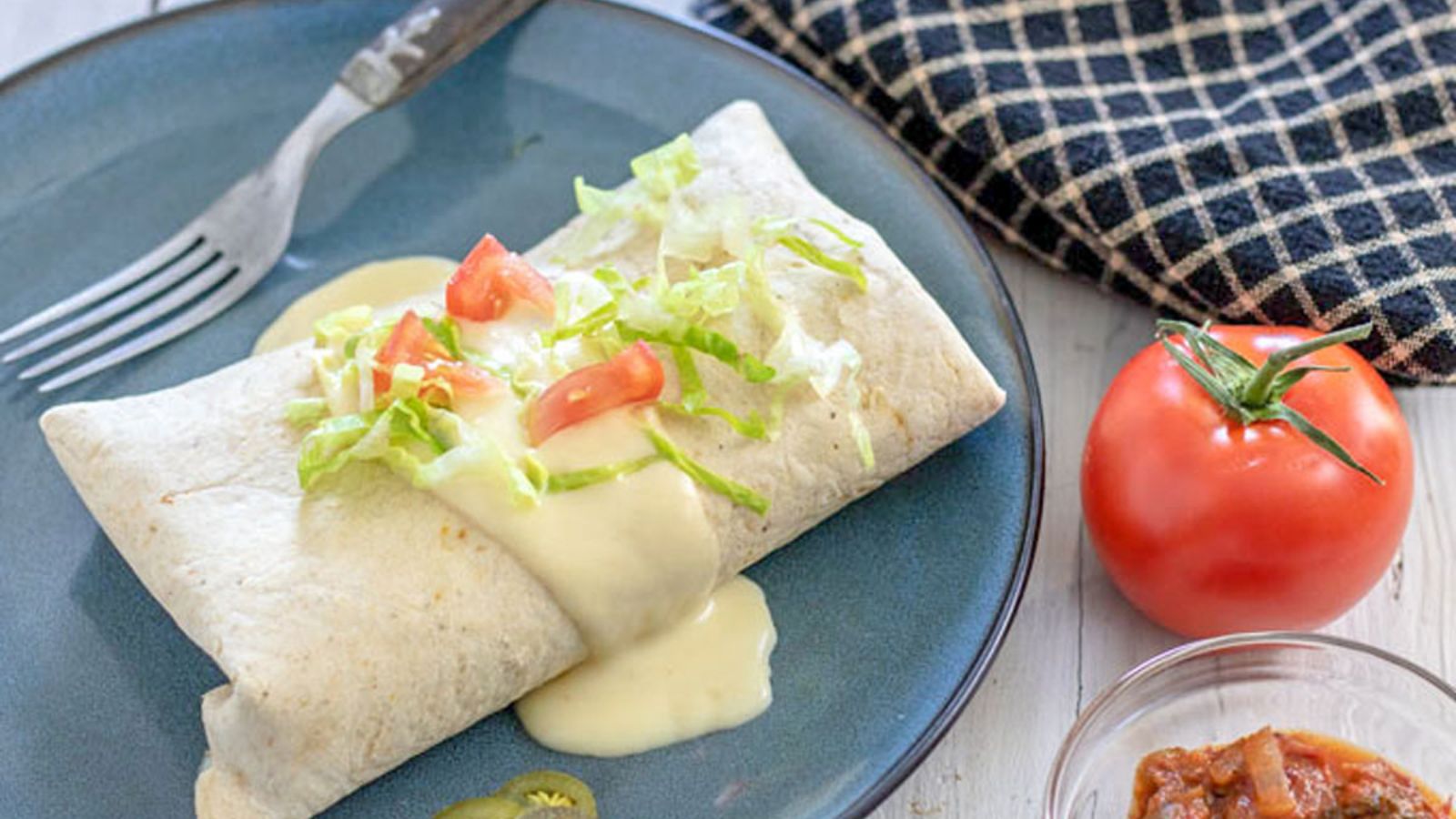 Burrito topped with cheese sauce and fresh tomatoes and lettuce on a blue plate.