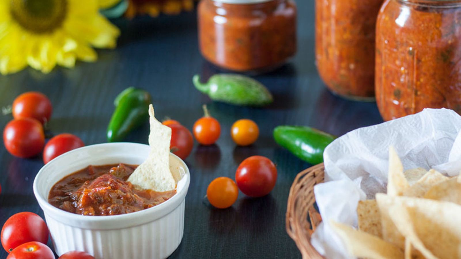 Bowl of smokey chipotle salsa with a tortilla chip in it surrounded by fresh tomatoes and peppers with jars of the salsa in the background.