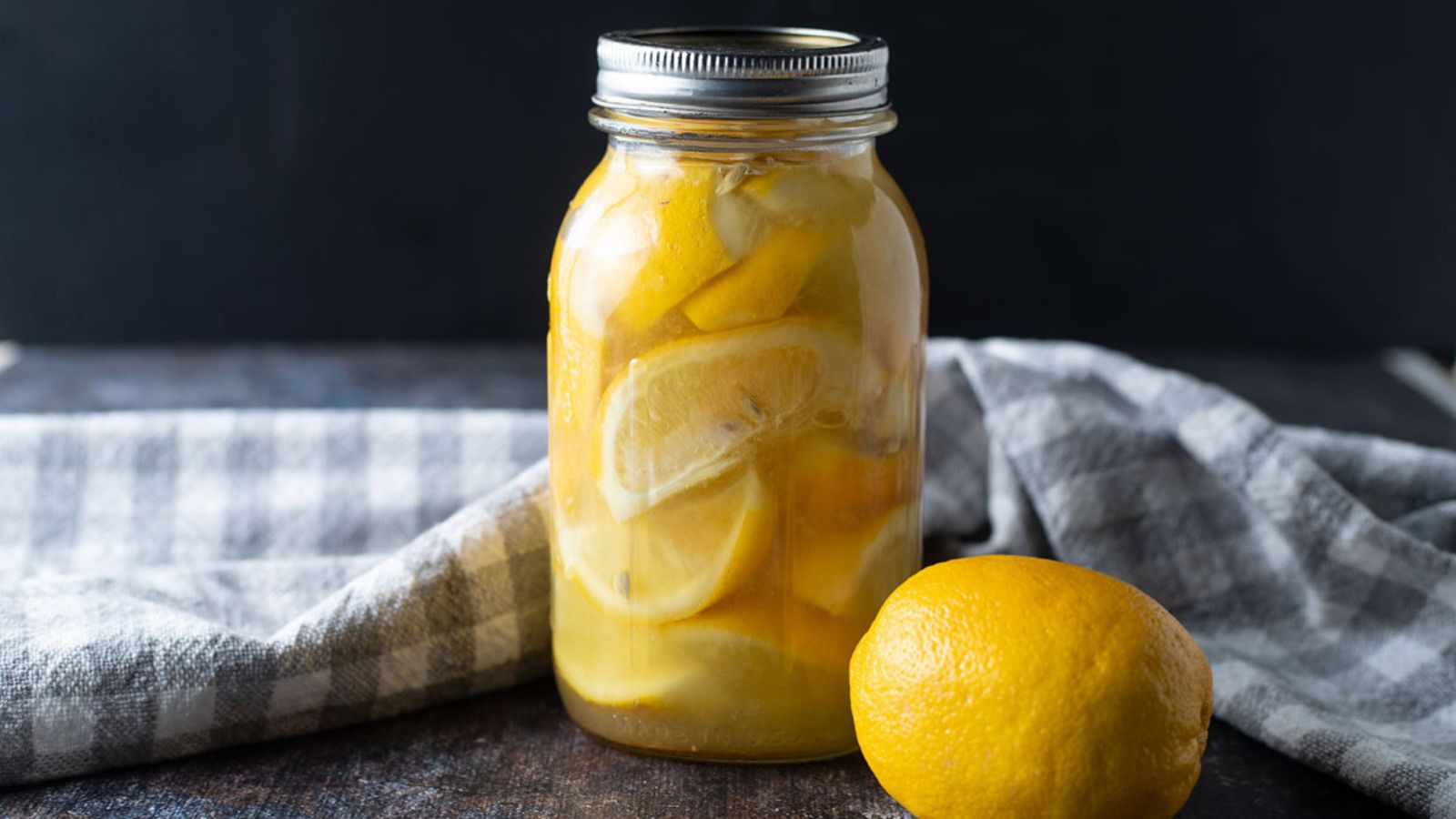 Jar of lemons with fresh lemon and gray checked tea towel.