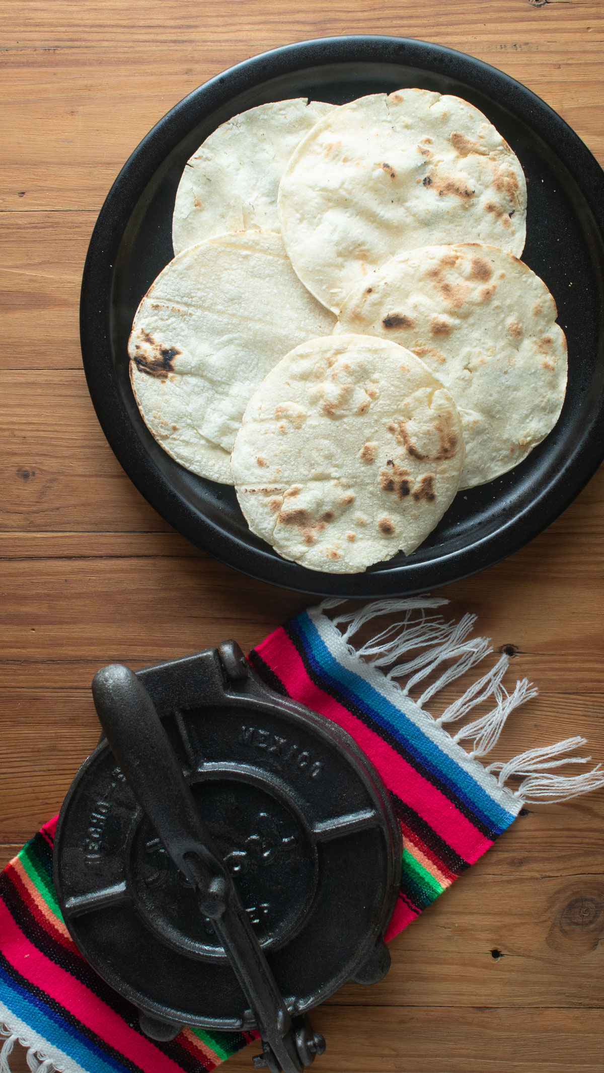 Five tortillas on a black plate with a tortilla press on a sarape.