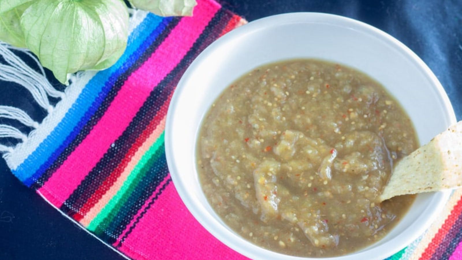 Corn chip dipped into green salsa which is in white bowl on a colorful Mexican blanket towel