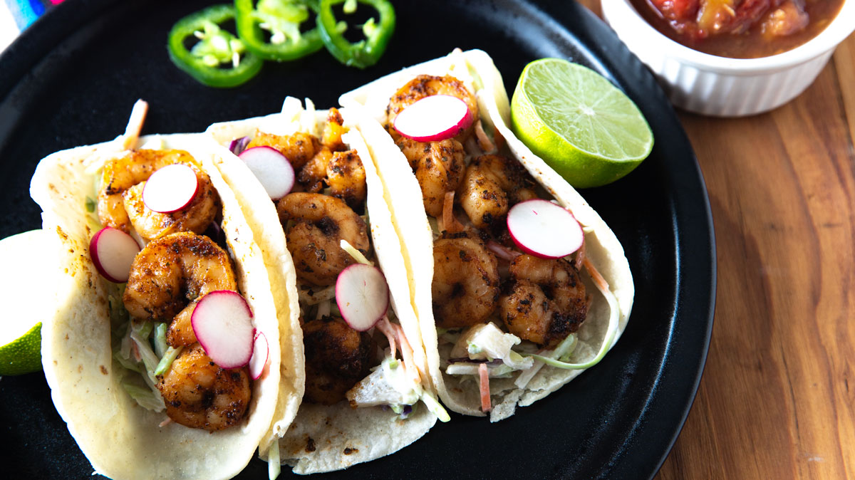 Three shrimp tacos on a black plate with sliced jalapenos.