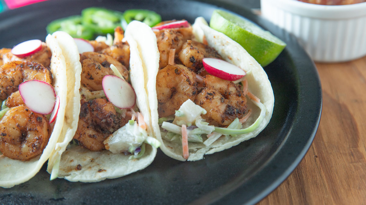 Three shrimp tacos on a black plate with lime and radish slices.