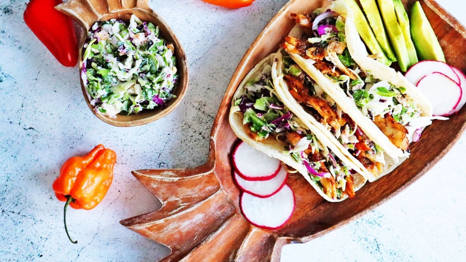 Three pieces of tacos on a plate with avocado slices and vegetables on the side.
