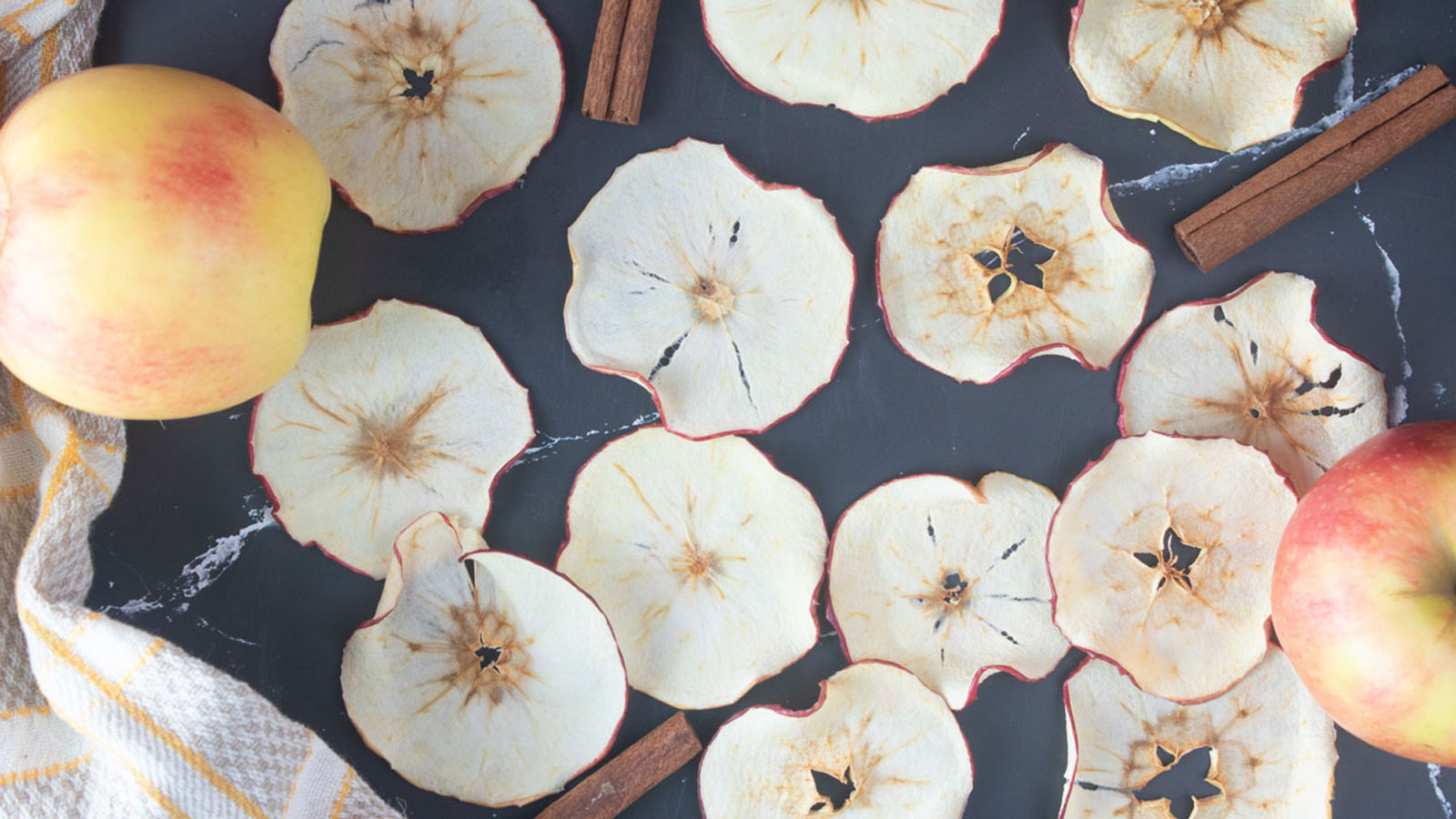 Dried apple slices on a dark board with cinnamon sticks.