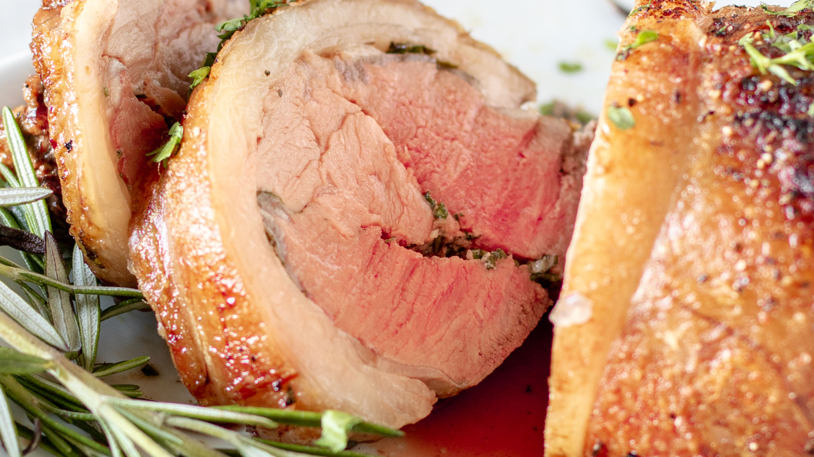 Sliced lamb loin, medium rare on white platter with fresh rosemary.