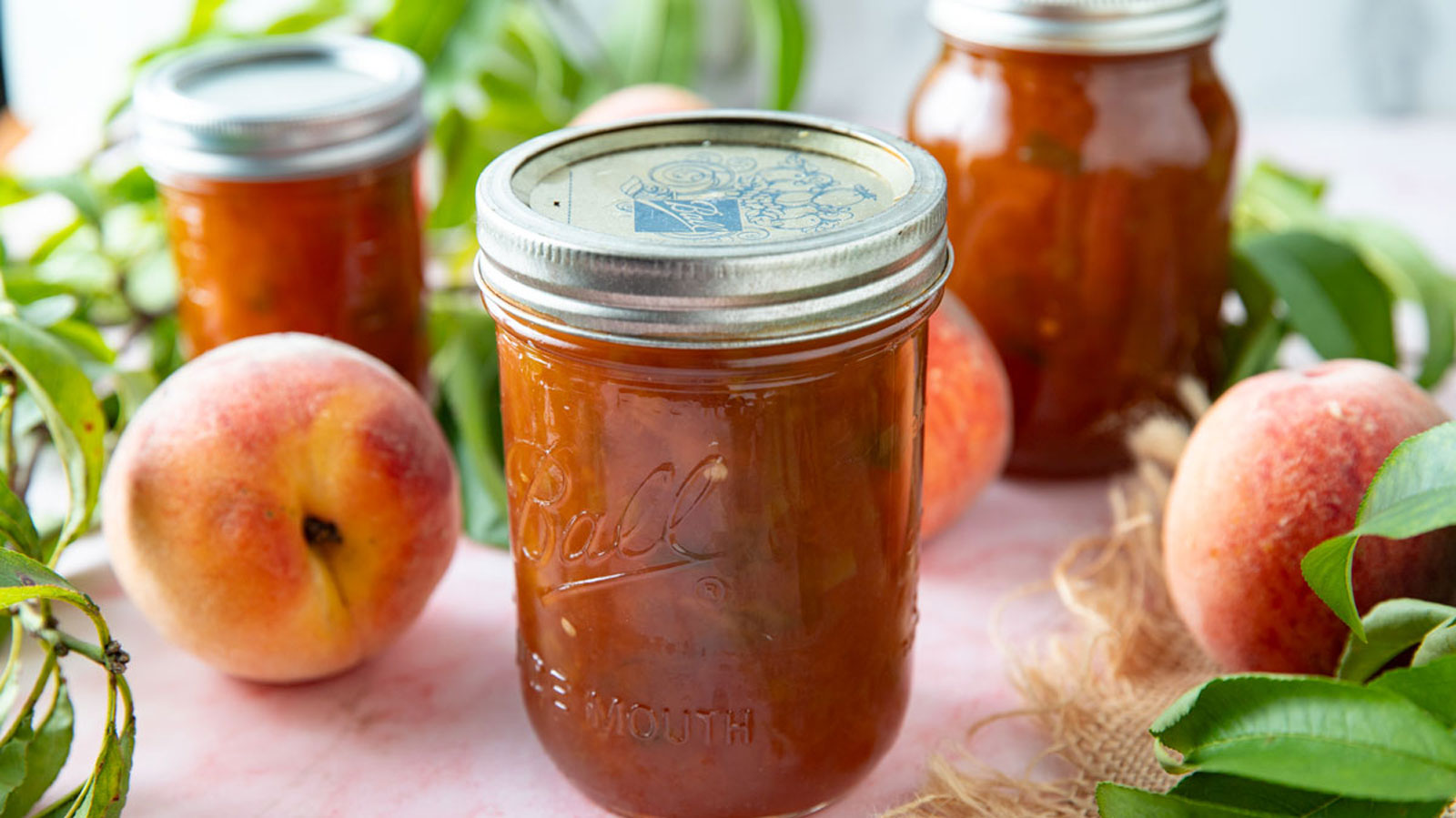 Jars of peach jalapeno jam on a pink board with fresh peaches.