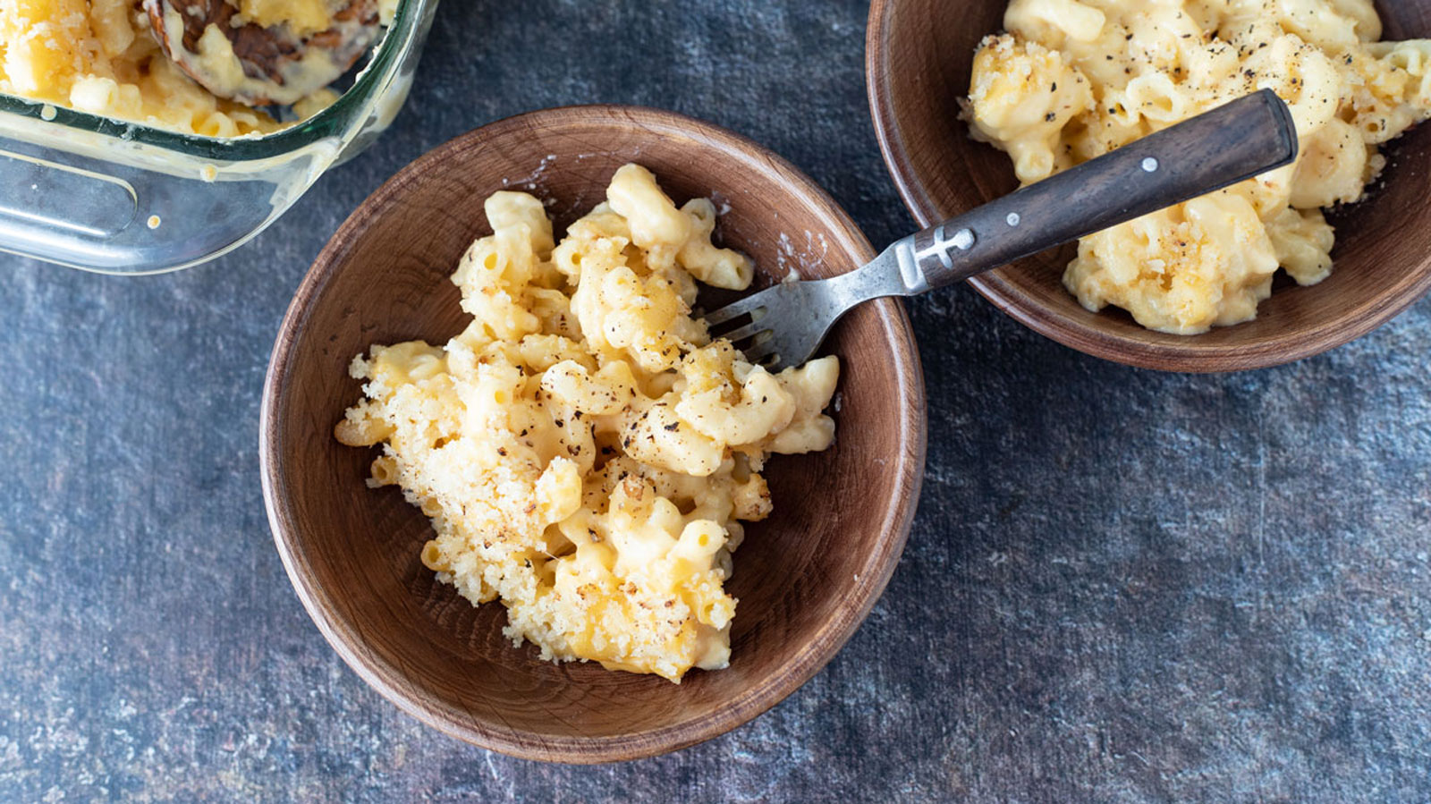Two wooden bowls of mac and cheese next to a casserole dish.