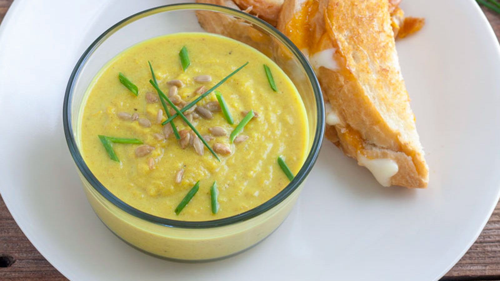 Yellow soup in a glass bowl with a grilled cheese sandwich on a white plate.