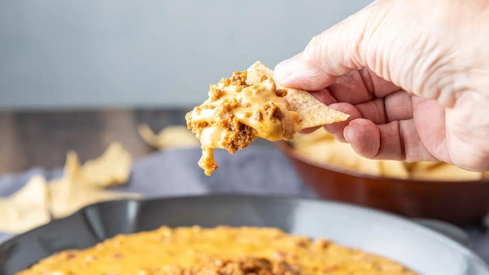 Woman's hand scooping chorizo dip out of bowl with a blue corn tortilla chip.