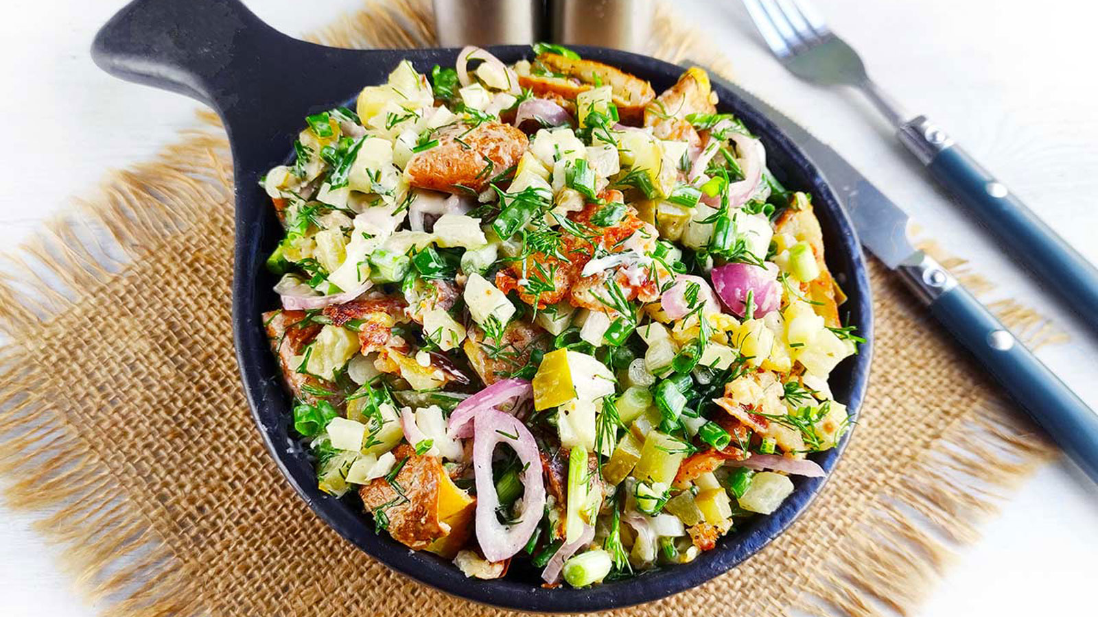 Colorful potato salad in a black bowl with utensils to the side.