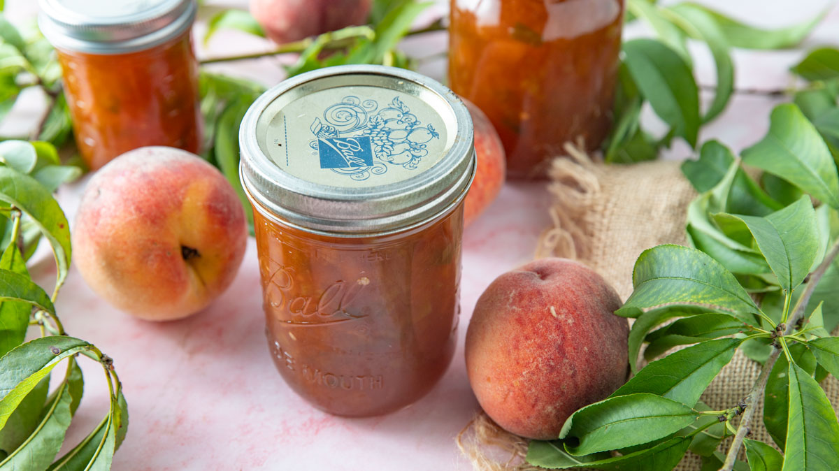 Jars of peach jam on a pink board with peach leaves.