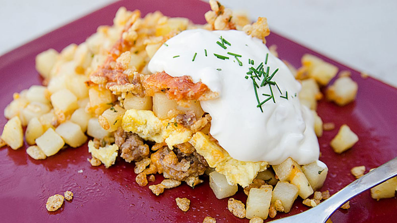 Pile of hash brown casserole on a red plate topped with sour cream.